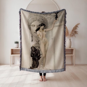 May include: A beige woven throw blanket featuring a classical art print. The artwork depicts a nude female figure with a large stone carving of a woman's face in the background. The blanket has a fringed edge.