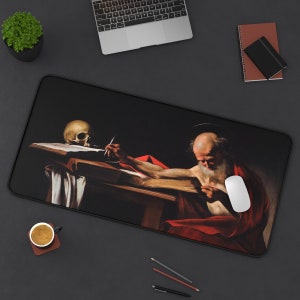 May include: A black and white mousepad with a painting of a man writing on a book with a skull on the table. The man is wearing a red robe and has a long white beard.