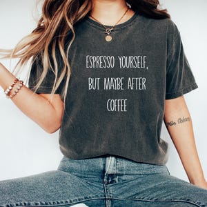 Può includere: T-shirt grigio scuro con la scritta bianca "Espresso Yourself, But Maybe After Coffee". La maglietta ha una vestibilità ampia e un aspetto vintage lavato. La modella indossa jeans blu.