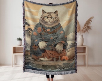 May include: Woven throw blanket featuring a large cat in a traditional Japanese style. The cat is depicted in a blue and orange kimono, with a serene expression. The background includes a sunset, mountains, and water.