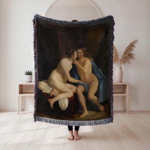 May include: Woven throw blanket featuring a classical painting of two nude figures in an embrace. The artwork has a dark background with a blue drape and a fringed edge. The blanket is held up by a person.