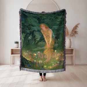 May include: A woven throw blanket displaying a classical painting of a woman in a forest. The artwork shows a woman in a golden dress surrounded by glowing lights and figures. The blanket has a dark green border with a fringed edge.