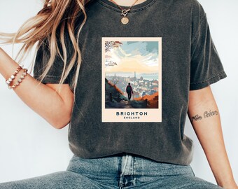 Könnte beinhalten: Ein dunkelgraues T-Shirt mit einer Grafik im Vintage-Stil, die eine Person zeigt, die auf einem Weg über eine Stadt geht, mit dem Text "Brighton, England".