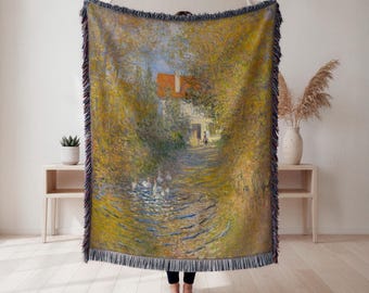 May include: A woven throw blanket displaying a landscape painting with a path, water, and a house with a red roof. The artwork uses a warm colour palette of yellows, greens, and blues, with a fringed edge.