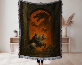 Op de afbeelding: Geweven sprei met een ridder te paard die een draak in een vurige boog tegenkomt. Het kunstwerk is in bruin-, zwart- en oranje tinten, met een gefranjerde rand. De deken wordt door een persoon omhoog gehouden.