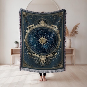 May include: A dark blue woven throw blanket featuring a celestial design with a central sunburst and surrounding constellations. The blanket has gold accents and a fringed edge. The design includes a detailed zodiac map.