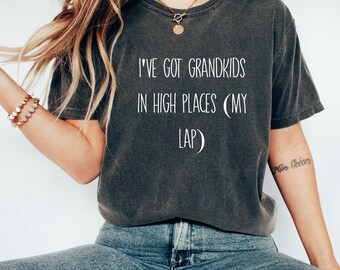 Puede incluir: Una camiseta gris oscuro con el texto "I've got grandkids in high places (my lap)" impreso en blanco. La camiseta presenta un gráfico de luna creciente.