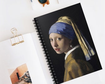May include: A spiral bound notebook with a black cover featuring a painting of a woman with a blue and yellow headscarf and a pearl earring. The woman is looking to the left of the image.