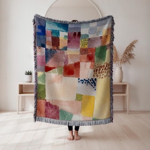 May include: A colourful woven throw blanket with a patchwork design. The blanket features a variety of colours including blue, red, yellow, green, and pink. The edges are finished with a fringe. The blanket is held up in front of a person.