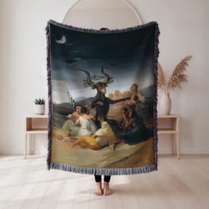 May include: A woven throw blanket featuring a dark, dramatic painting. The artwork depicts a goat-like figure surrounded by people in a landscape under a night sky with a crescent moon. The blanket has a fringed edge and is held up by a person.