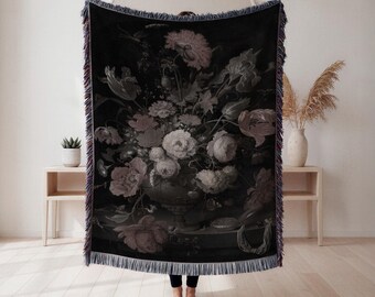 May include: Black throw blanket with a floral still life design in pink, grey, and white. The blanket has a fringed edge with mixed colours. The artwork shows a vase overflowing with flowers.