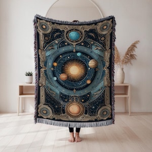 May include: A woven tapestry blanket featuring a celestial design with a central sun, planets, and stars in shades of blue, gold, and brown. The blanket has a decorative border and fringe.