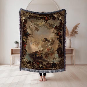 May include: Woven throw blanket featuring a classical painting with figures in a cloudy sky. The blanket has a dark border with a fringe. The color palette includes beige, brown, blue, and red.