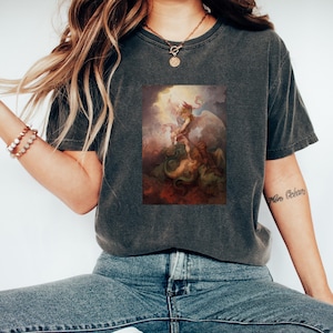 May include: A dark gray t-shirt with a vintage-style print of an angel battling a dragon. The print is in shades of brown, red, and yellow.