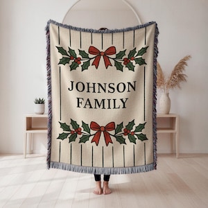 May include: A beige woven throw blanket with a black vertical stripe pattern and a decorative border. The blanket features the text "JOHNSON FAMILY" in a rectangular frame, with holly and red bow accents. The edges have a blue fringe.