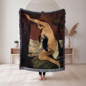 May include: Woven throw blanket featuring a classical painting of a nude figure in a bedroom setting. The artwork depicts a woman with dark hair, draped in fabric, against a backdrop of red and green tones. The blanket has a fringed edge.