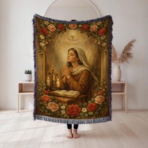 May include: A woven tapestry throw blanket featuring a religious painting of a woman praying, surrounded by roses. The artwork includes a cross, candles, and an open book. The blanket has a fringed edge and is predominantly beige, brown, and red.