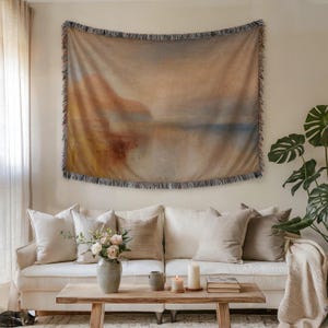 Könnte beinhalten: Ein gewebtes Wandbehang mit einem Landschaftsdesign in Braun-, Beige- und Blautönen hängt über einem weißen Sofa. Der Wandteppich hat einen Fransenrand. Der Raum ist mit neutralfarbenen Kissen, einem Holztisch und einer Topfpflanze dekoriert.