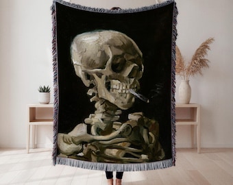 Puede incluir: Manta tejida negra con una pintura de un esqueleto fumando un cigarrillo. La obra de arte utiliza tonos beige, marrón y negro, sobre un fondo oscuro. La manta tiene un borde con flecos.
