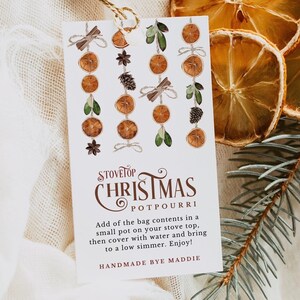 May include: A white card with a watercolor illustration of a stovetop Christmas potpourri. The card has the text "Stovetop Christmas Potpourri" and instructions for using the potpourri. The card is sitting on a white fabric background with dried orange slices and a pine branch.