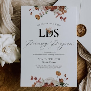 Könnte beinhalten: Eine weiße Einladungskarte mit floralen Akzenten und dem Text "LDS Primary Program" liegt neben einem weißen Umschlag mit einem goldenen Siegel. Die Karte enthält das Datum "10. November" und die Uhrzeit "9-10 Uhr". Die Artikel sind auf einer Holzoberfläche angeordnet.