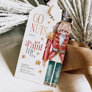 May include: A Christmas gift tag featuring a watercolour illustration of a nutcracker. The tag reads "Go Nuts and Paint Me" with a star and a happy holidays message.