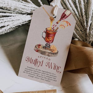 Könnte beinhalten: Ein rechteckiges Geschenk-Etikett mit einer Aquarell-Illustration von Glühwein, der in ein Glas gegossen wird. Das Etikett trägt die Worte "Spiced Mulled Wine" und Anweisungen. Das Etikett ist mit einer weißen Schnur gebunden.