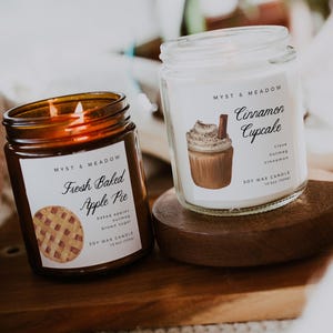 Puede incluir: Dos velas de cera de soja en frascos de vidrio sobre una bandeja de madera. Una vela está etiquetada como "Fresh Baked Apple Pie" y la otra como "Cinnamon Cupcake". Las etiquetas incluyen ilustraciones de los aromas. Las velas son de "Myst & Meadow".