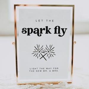 Könnte beinhalten: Ein goldfarbener Rahmen mit dem Text "LET THE spark fly" in schwarzer Fettschrift. Unter dem Text befindet sich eine Schwarzweißillustration von zwei gekreuzten Wunderkerzen. Der Text "LIGHT THE WAY FOR THE NEW MR. & MRS." befindet sich unter der Illustration.