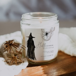May include: A white candle jar with a black and white illustration of a witch and a spider web. The label reads "Halloween Darkness" and "Hand poured in Seattle 10.5 oz (300 g) Whiskey Cedar Co."