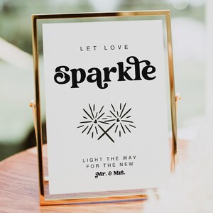 Könnte beinhalten: Ein goldfarbener Rahmen mit dem Text "Let love sparkle" in Schwarz, der ein stilisiertes Bild von zwei Funken mit Herzen zeigt. Der Text darunter lautet "Light the way for the new Mr. & Mrs."