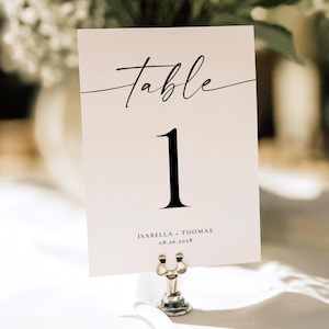 May include: A rectangular white table number card held by a small silver metal stand. The card features the word Table in black script, the number 1 in a large serif font, and the text ISABELLA + THOMAS 08.26.2028 printed in black at the bottom.