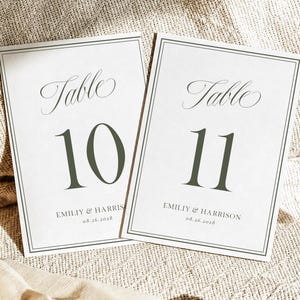 May include: Two rectangular white table cards with a thin dark gray border. Each card features the word Table in elegant script, the numbers 10 and 11 in a large dark gray serif font, and the text EMILIY & HARRISON with the date 08.26.2028 below.