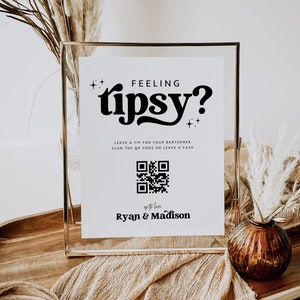 May include: A white sign with black text that reads "Feeling Tipsy?" with a QR code and the text "Leave a tip for your bartender, scan the QR code or leave a cash. With love, Ryan & Madison."