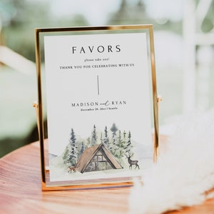 May include: A white sign with gold trim in a gold frame on a wooden table. The sign says "Favors, please take one! Thank you for celebrating with us, Madison and Ryan, December 29, 20xx, Seattle". The sign has a watercolor illustration of a cabin in the woods with deer.