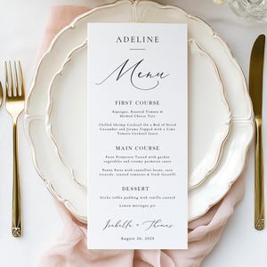 May include: A white rectangular menu card with black serif and script text centered on a white scalloped plate. The menu lists courses for Adeline, Isabella, and Thomas on August 26, 2028. Gold-toned cutlery sits on either side of the plate.
