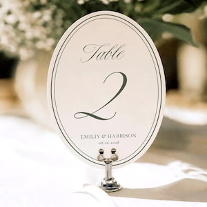 May include: An oval-shaped table number card featuring a cream background with dark gray double-line borders. The text reads Table 2, Emiliy & Harrison, and 08.26.2028. The card is held upright by a small, silver-toned metal stand with a circular base.