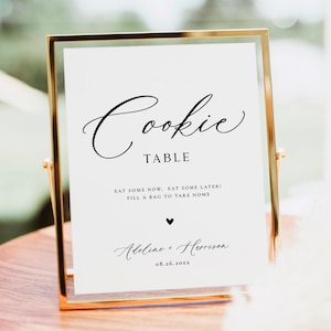 May include: A gold-framed sign with the words "Cookie Table" in elegant script. Below, it reads "Eat some now, eat some later! Fill a bag to take home." The sign includes a heart and the names "Adeline + Harrison" with a date.
