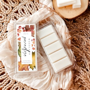 Puede incluir: Un derretimiento de cera de soja de seis cubos en un recipiente de plástico transparente. El derretimiento de cera es blanco y el recipiente tiene una etiqueta con un diseño de acuarela de hojas de otoño y el texto "WOODLAND paradise Whiskey Cedar Co. SOY WAX MELT Hand poured in Seattle 1.4 oz (40 g)"