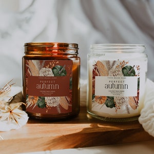 Puede incluir: Dos tarros de velas de vidrio con etiquetas que dicen "Perfect Autumn" y que presentan un diseño floral con calabazas, maíz y trigo. Las velas están sobre una superficie de madera.