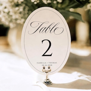 May include: An oval-shaped white table number card featuring a thin black border, the word Table in elegant script, the number 2 in a bold serif font, and the text ISABELLA + THOMAS 08.26.2028, held upright by a small silver metal stand.