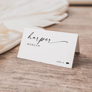 May include: A white folded place card featuring the name Harper in a black cursive script font, with the name MORGAN printed in a black sans-serif font below it. The bottom right corner displays the text table 6 and a small black pig icon.
