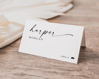 May include: A white folded place card featuring the name Harper in a black cursive script font, with the name MORGAN printed in a black sans-serif font below it. The bottom right corner displays the text table 6 and a small black pig icon.