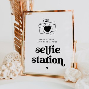 May include: A gold-framed sign with the words "selfie station" in a bold, black font. The sign features a camera icon with a heart in the lens and the text "Grab a prop and take a pose."