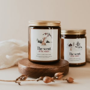 May include: Two amber glass soy candles with gold lids and labels. The labels feature illustrations of a fox and a house in a snowy winter scene. The text on the labels reads "The scent of the winter" and "Frosty Flame Creations".