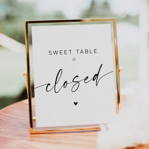 May include: A gold-framed sign with the text "SWEET TABLE IS closed" in black script. A small black heart is below the word "closed". The sign is displayed on a wooden table.