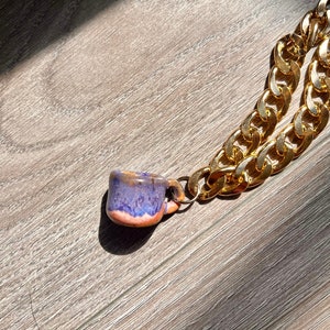 May include: A gold chain necklace with a small ceramic coffee cup pendant. The cup is glazed with shades of purple, orange, and brown. The chain has large, interlocking links.