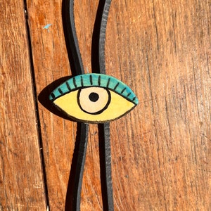 May include: A handmade bolo tie featuring a painted eye pendant. The eye is yellow with a black pupil and blue eyelashes. The tie is made of black leather and rests on a wooden surface.