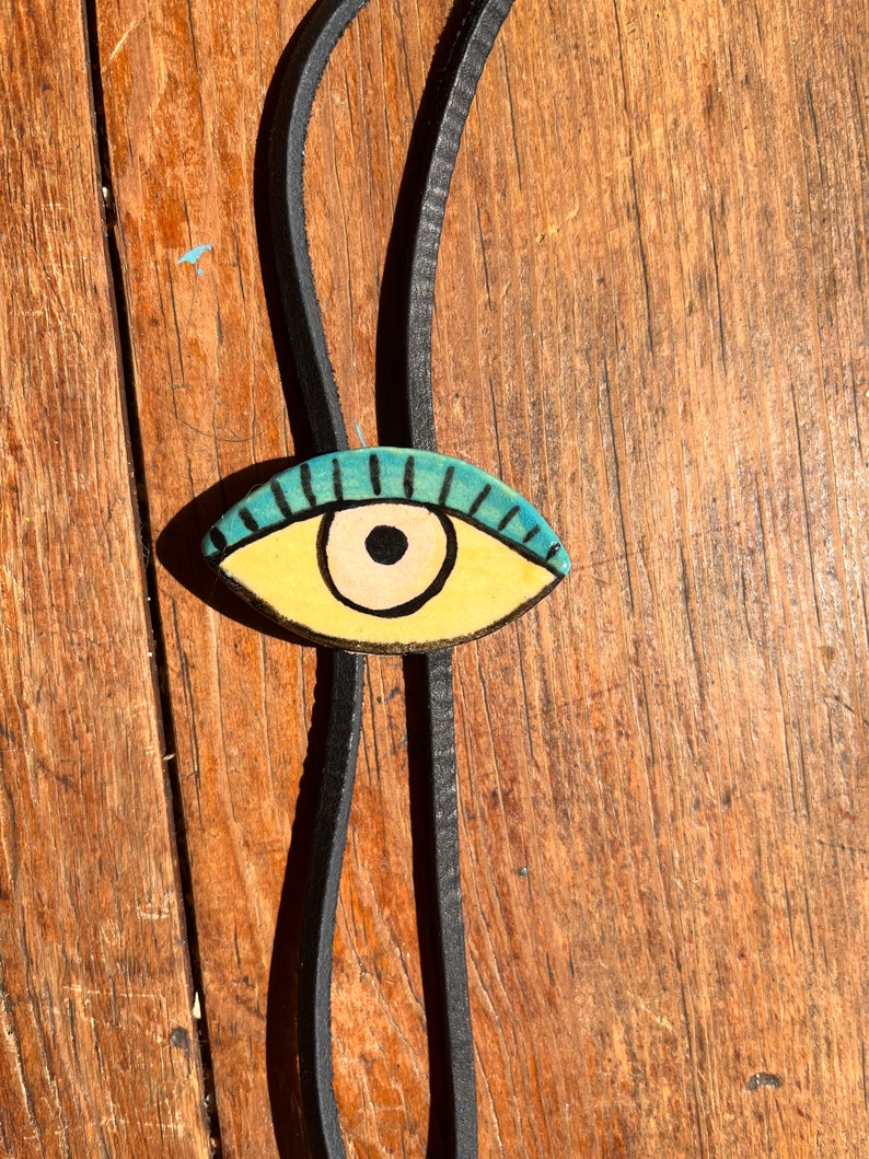 May include: A handmade bolo tie featuring a painted eye pendant. The eye is yellow with a black pupil and blue eyelashes. The tie is made of black leather and rests on a wooden surface.