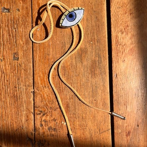 May include: A bolo tie featuring an eye-shaped pendant with a blue iris, black outline, and tan-colored lashes. The pendant is attached to a tan leather cord with silver-colored metal tips. The tie rests on a wooden surface.